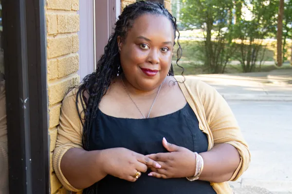 A black woman with braids in a black dress and yellow cardigan smiles at the camera